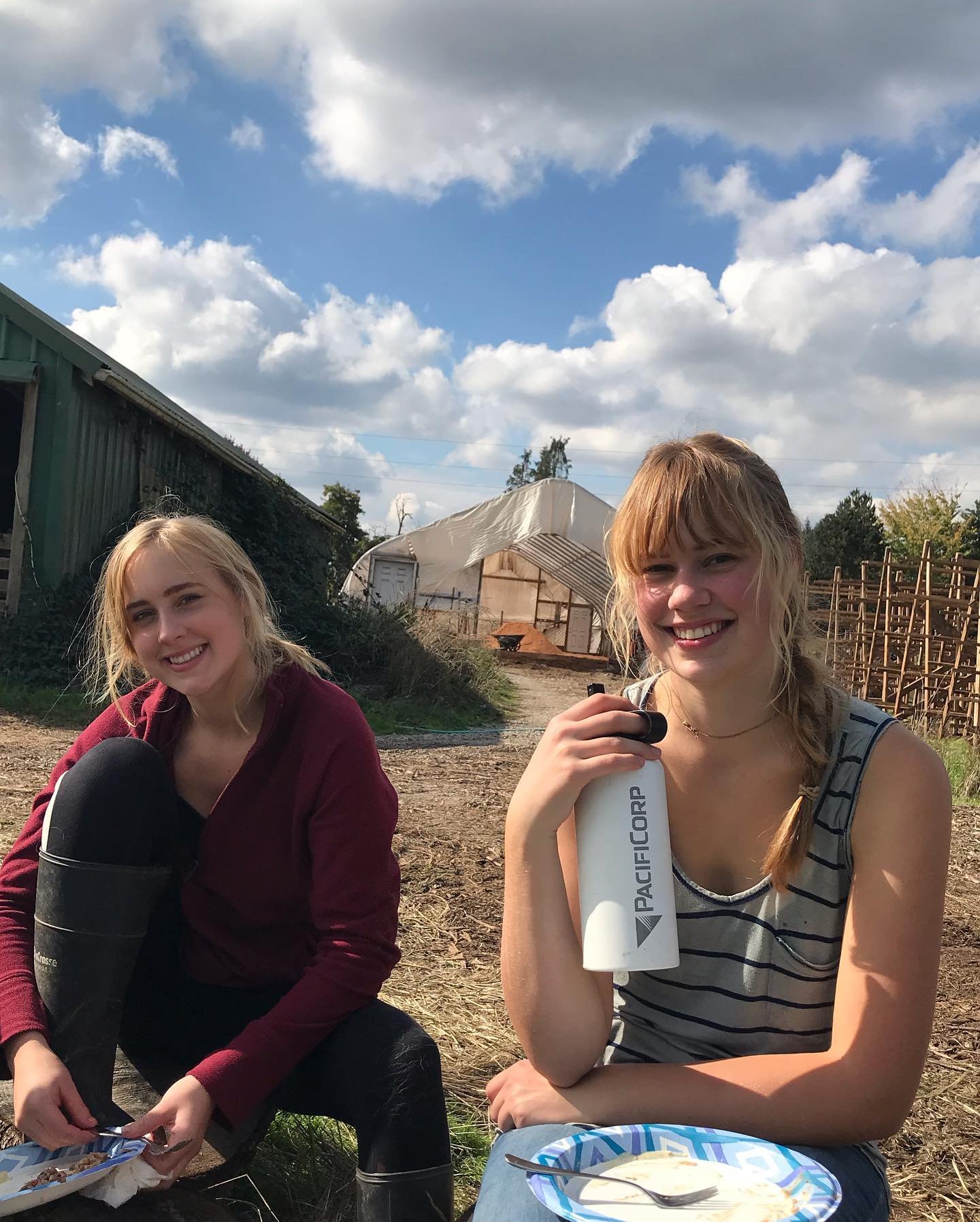 475842100_9086259698075746_136586530656458843_n Two young women enjoying a meal outdoors on a farm, with one holding a water bottle and the other preparing food. The background features a farm structure and blue sky with clouds, highlighting a relaxed, rural atmosphere.