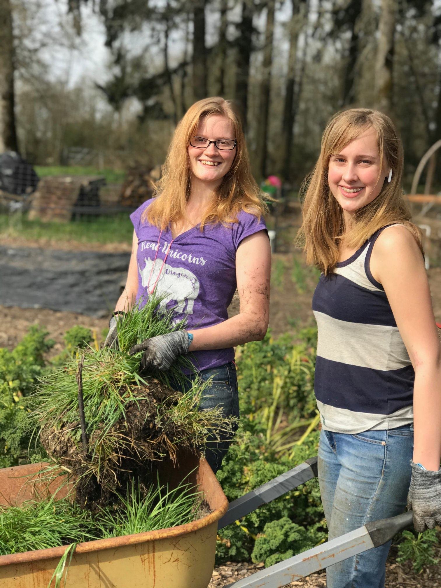 90440992_2785980311437081_1195611250441060352_n Two young women smiling while gardening, one holding a wheelbarrow filled with freshly pulled grass and weeds. They are wearing gloves and casual clothing, surrounded by lush greenery in a community garden setting.