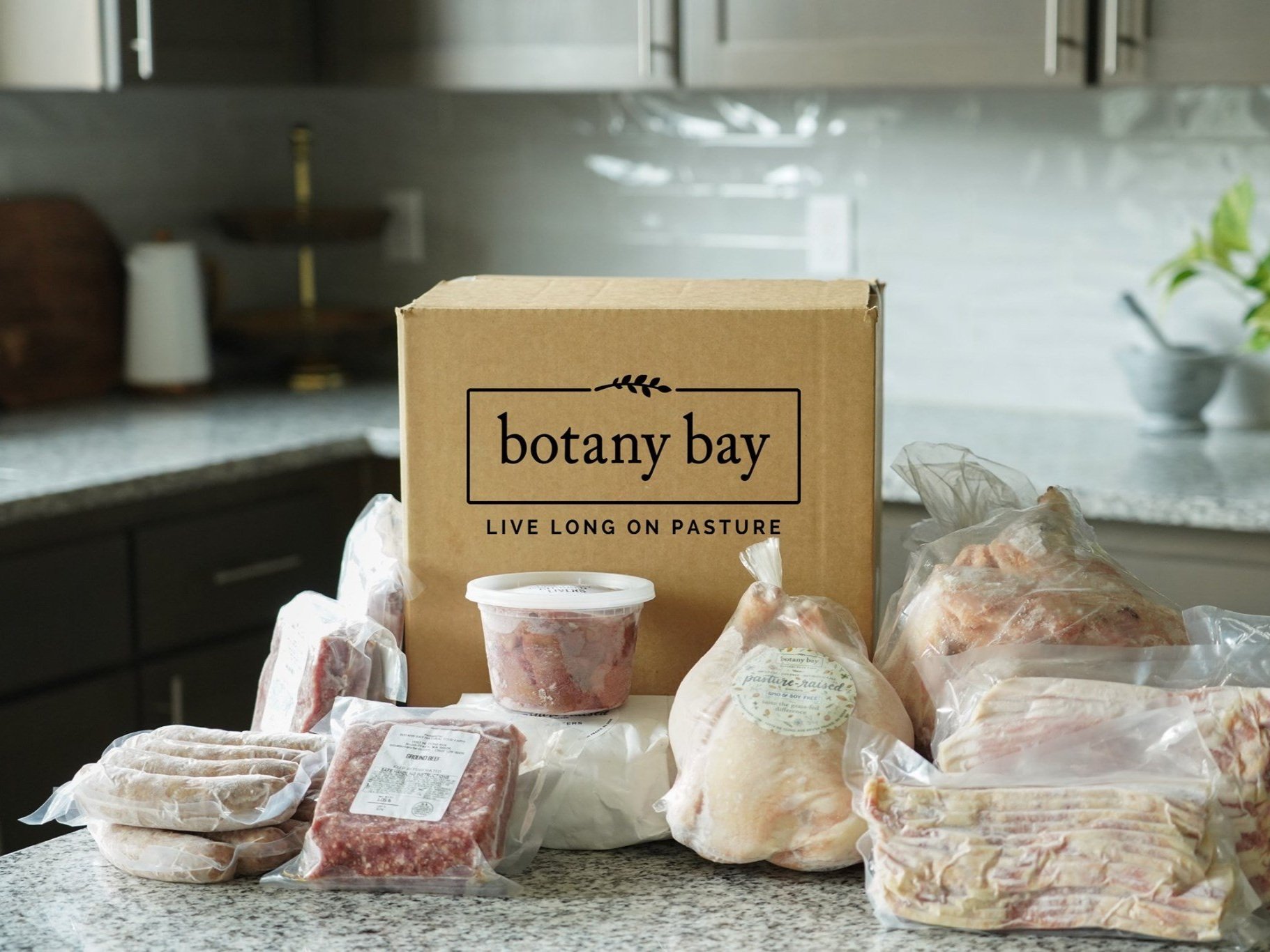 Box of Botany Bay pasture-raised meats and products displayed on a kitchen countertop, featuring various cuts of chicken, sausages, ground meat, and bacon, emphasizing quality and sustainability in food sourcing.