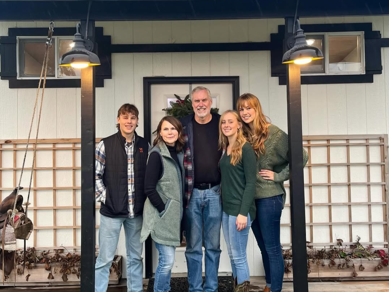 Family portrait in front of a cozy house, featuring five individuals smiling together. The group includes three young adults and two middle-aged adults, with a festive wreath visible on the door. The setting showcases outdoor lighting and a wooden trellis, creating a warm and inviting atmosphere.